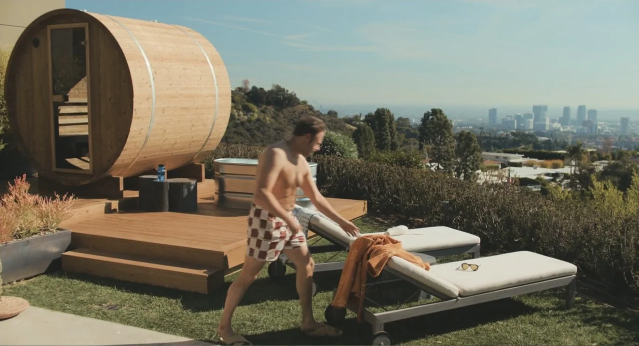 Good Fortune (2025) movie still: fullbody — A wooden barrel sauna sits on a deck overlooking a distant city skyline. A man wearing swim trunks walks…