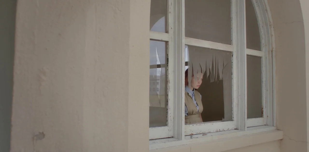 Darlin’ (2019) movie still: medium — A light-colored building facade features an arched window with several shattered, jagged glass panes. A young…