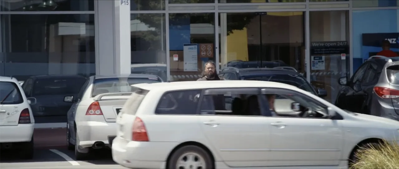 Bludgeon (2018) movie still: medium — A man stands in an outdoor parking lot in front of a glass building. Several cars are parked in rows while a…