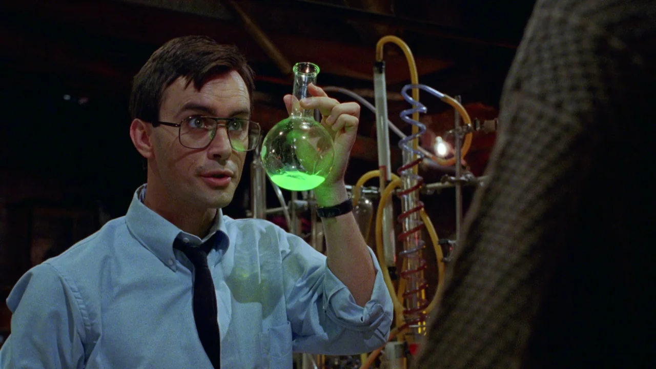 Bride of Reanimator (1989) movie still: medium — A dimly lit room filled with complex glass tubes and distillation equipment. A man holding glasses…