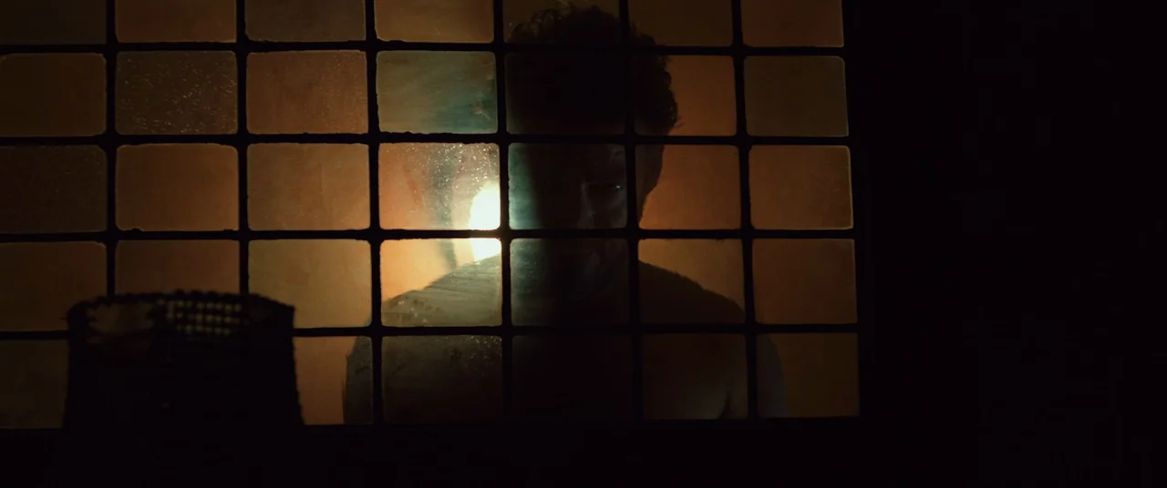 Speak No Evil (2022) movie still: medium — A dark space viewed through a grid of frosted glass panes illuminated from behind. A man's face and…