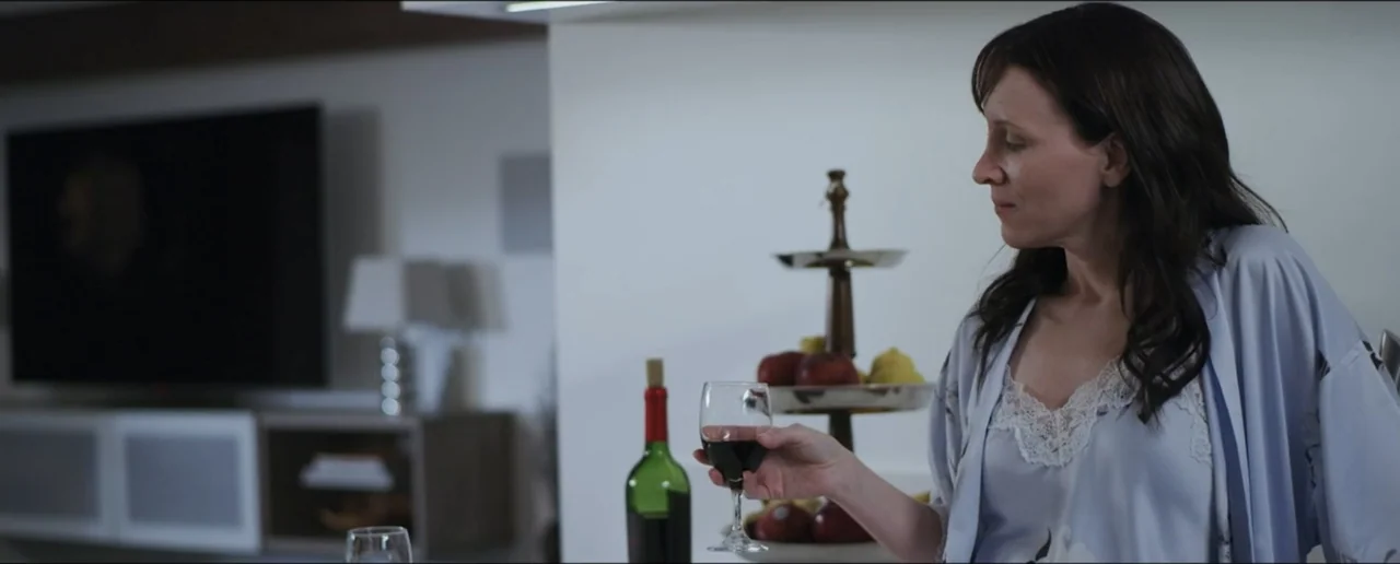 Hostages (2017) movie still: medium — A modern home interior features a kitchen counter with a wine bottle and fruit. A middle-aged woman in a light…