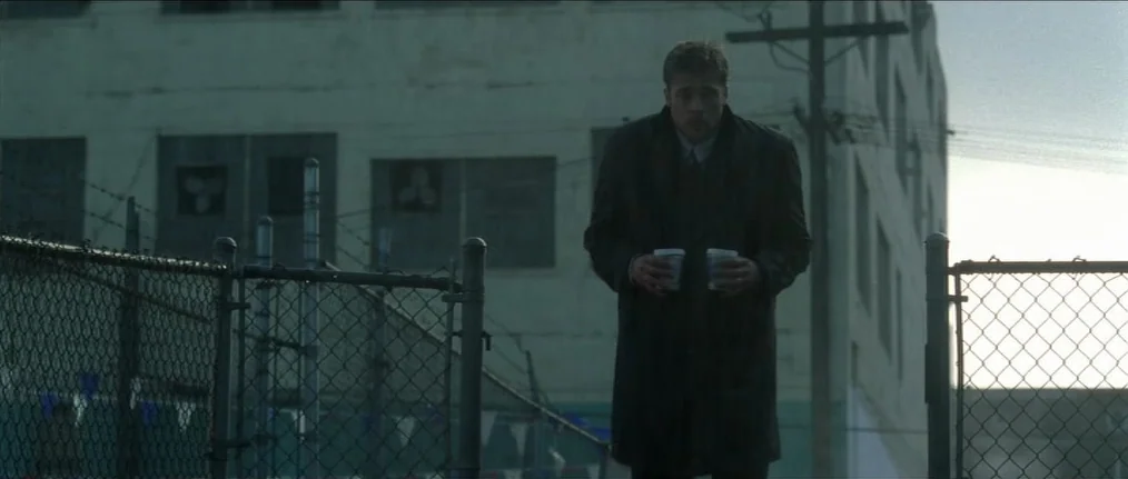 Se7en (1995) movie still: fullbody — An industrial lot features a chain-link fence in the foreground against a light-colored building. A man walks…
