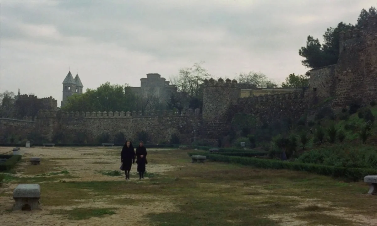 Tristana (1970) movie still: wide — A wide dirt courtyard is enclosed by massive medieval stone fortification walls and towers. Two figures dressed…