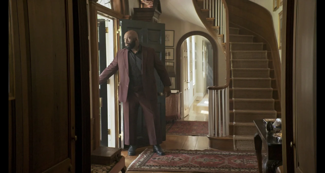 The Poison Rose (2019) movie still: fullbody — A formal home foyer features a sweeping staircase and detailed patterned rugs. A man in a suit stands…