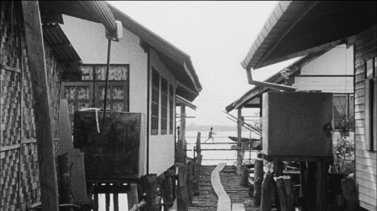 Mysterious Object at Noon (2000) movie still: wide — A narrow wooden walkway passes between houses built on stilts over water. In the distant…