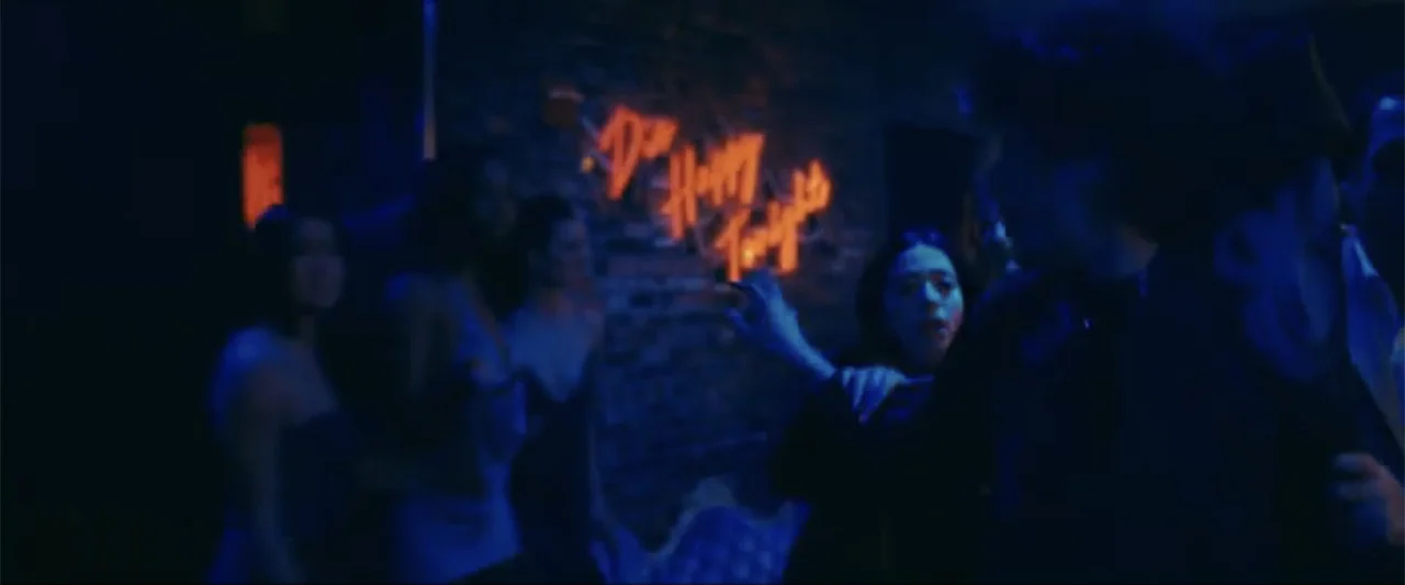 Anora (2024) movie still: medium — A crowded dimly lit nightclub interior with a brick wall behind a neon sign. Multiple people are moving in the…