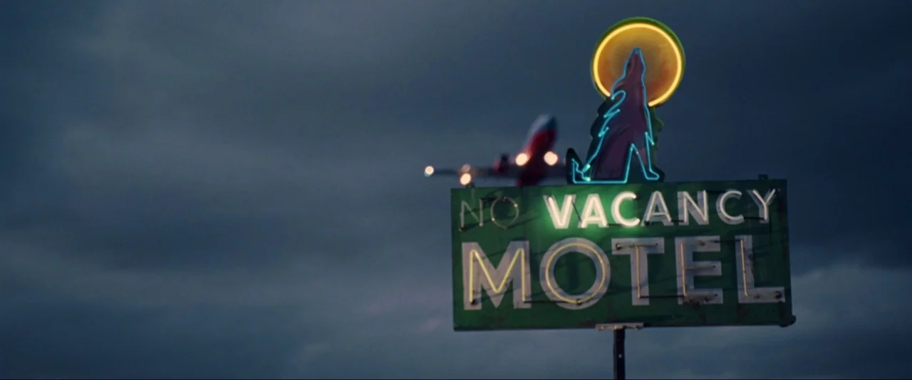 Kiss Kiss Bang Bang (2005) movie still: wide — A green neon motel sign stands against a dark, overcast night sky. An out of focus airplane flies…