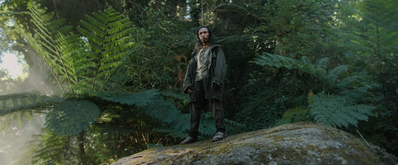 Boy Kills World (2023) movie still: fullbody — A dense, humid-looking environment filled with large, spreading tree ferns and mossy vegetation. A…