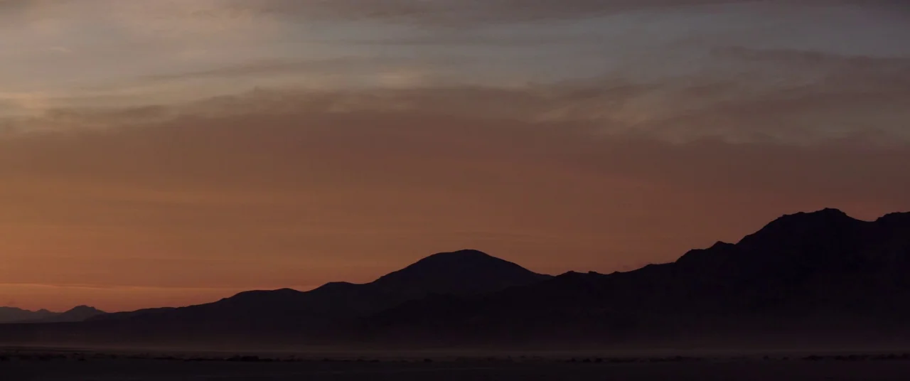 The Bad Batch (2016) movie still: establishing — A vast desert expanse stretches out beneath a range of dark mountain silhouettes. The sky displays…