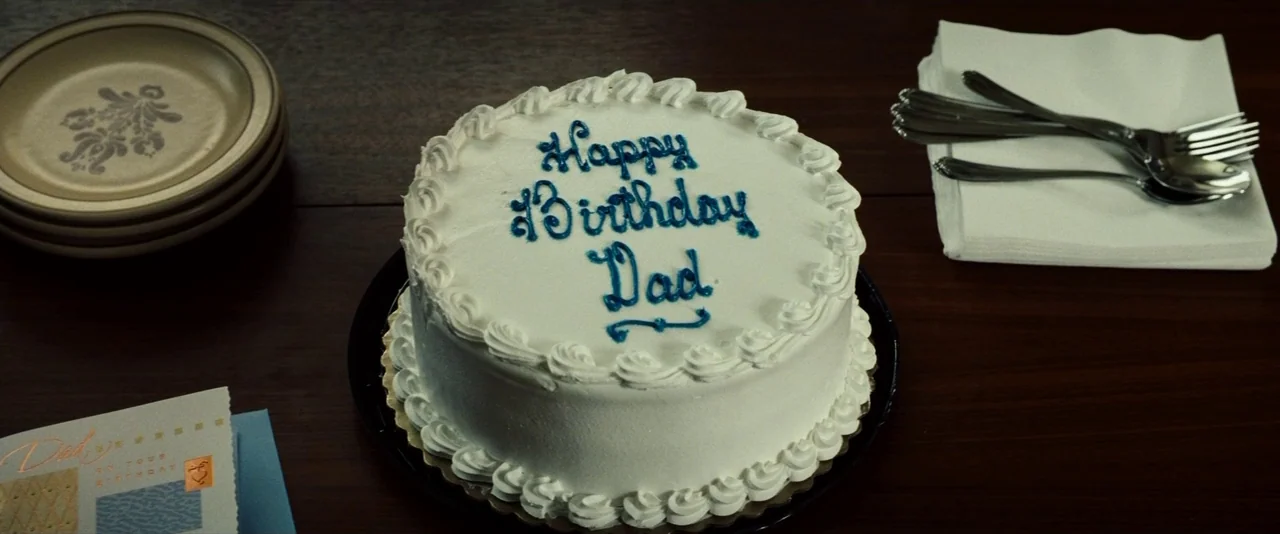 Gran Torino (2008) movie still: closeup — A frosted cake with blue icing sits on a dark wooden table surface. A stack of small plates, a greeting…