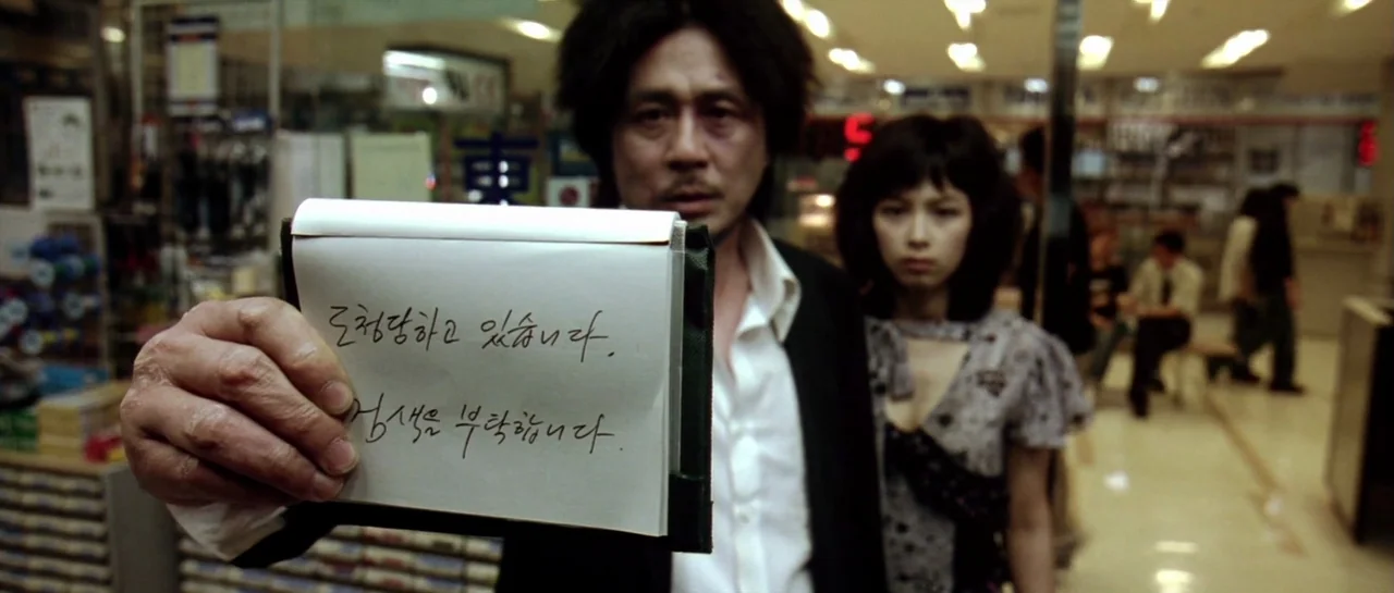 Oldboy (2003) movie still: medium — A crowded convenience store with shelves of products in the blurred background. A disheveled man holds a written…