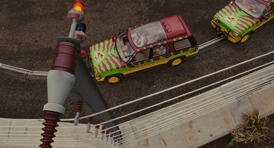 Jurassic Park (1993) movie still: wide — A high-angle view shows two marked SUVs driving along a rail track. An electric power pole stands in the…