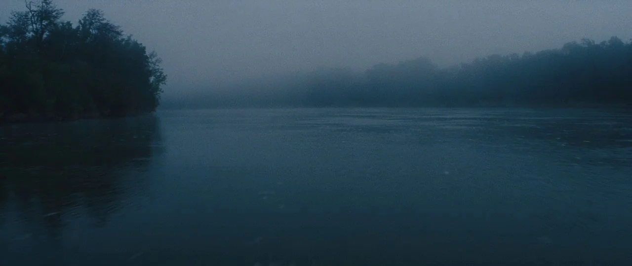 Mud (2012) movie still: establishing — A wide, still river flows between densely forested banks under a heavy, low-hanging fog. The water surface is…