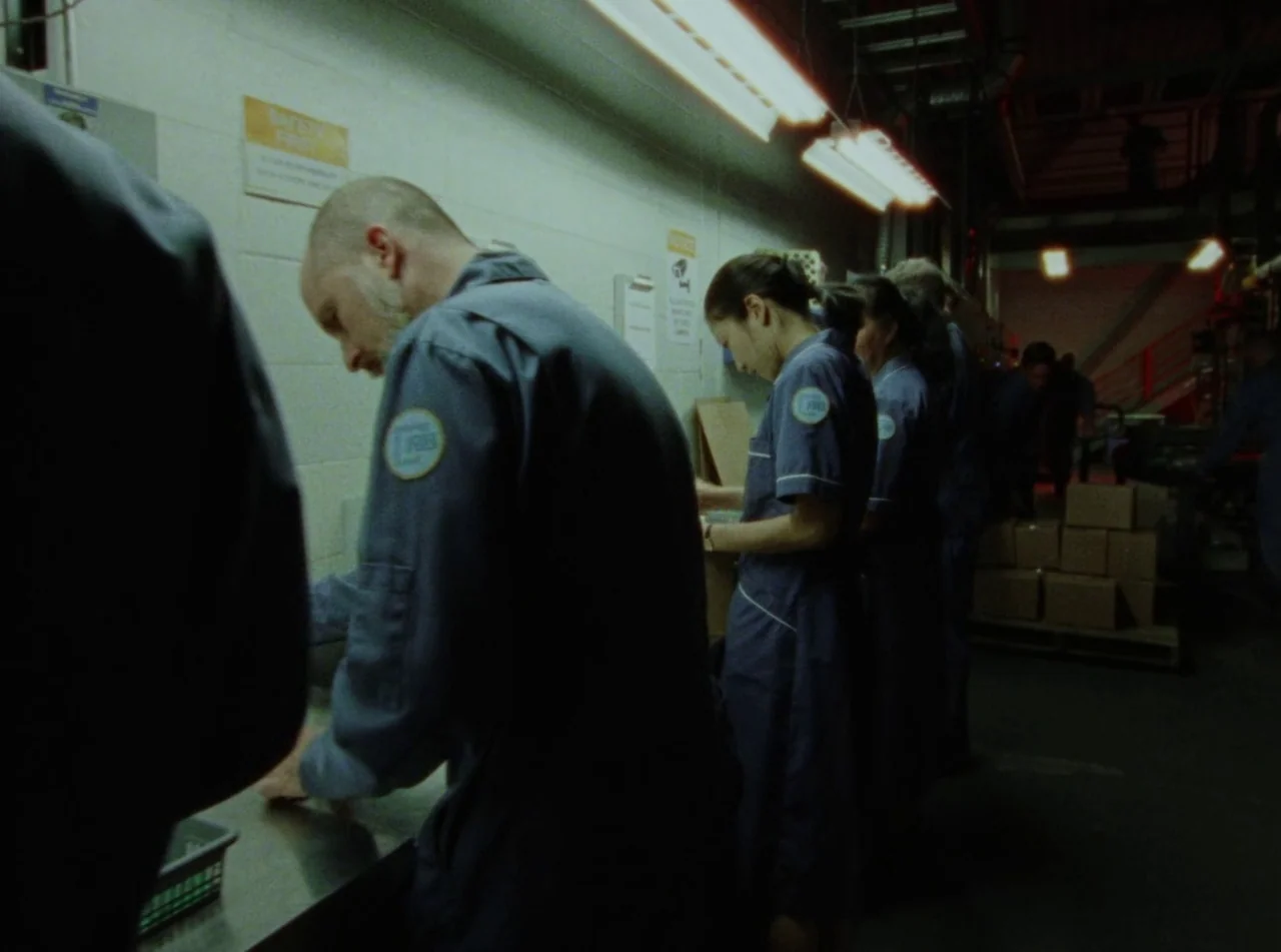 Riceboy Sleeps (2022) movie still: medium — An industrial workspace features long rows of stainless steel tables under bright fluorescent lighting.…