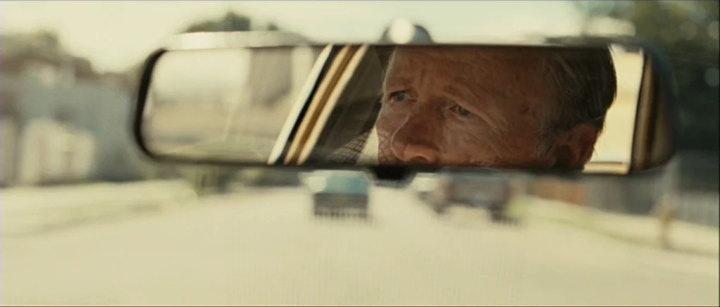 No Country For Old Men movie still: closeup — The interior of a vehicle seen through the rearview mirror. An elderly man with grey hair is reflected…