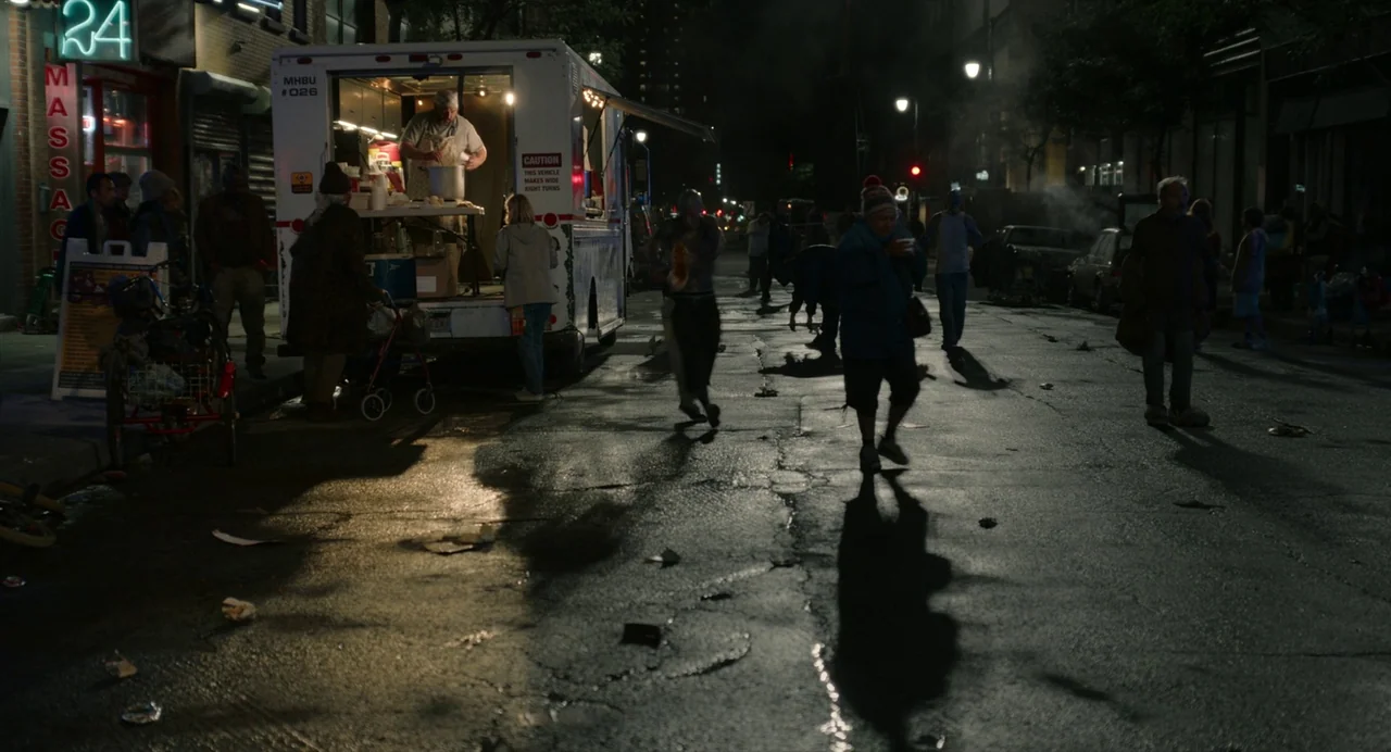 Beau Is Afraid (2023) movie still: wide — An urban night scene shows people passing a food truck on a damp pavement. The street is dimly lit by…