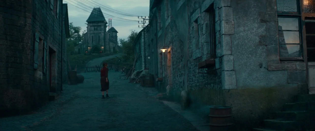 Overlord (2018) movie still: wide — A quiet cobblestone street winds through a stone village toward a distant stone church. A lone woman walks away…