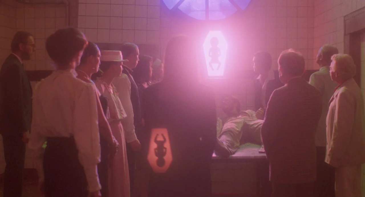 The Spider Labyrinth (1988) movie still: wide — A group of people stands in a tiled room with a glowing pink fetus icon. A body lies on a metal…