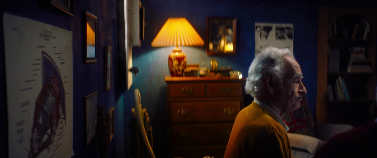 Linoleum (2022) movie still: medium — A dimly lit room with deep blue walls features a wooden dresser and a bookshelf. An elderly man in a yellow…