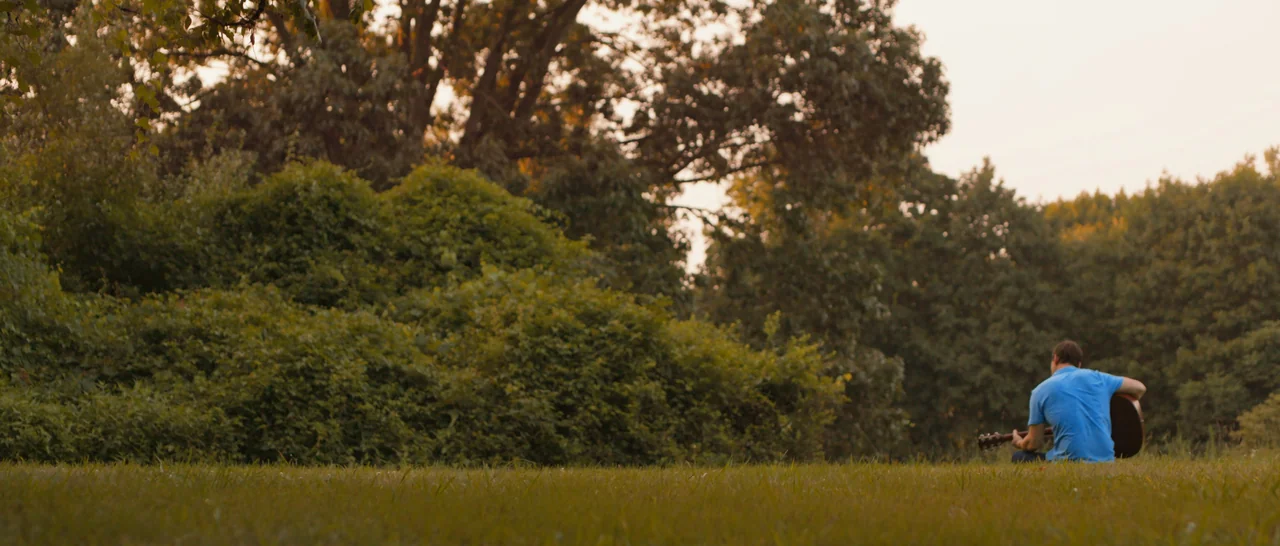 Songs For A Sloth (2021) movie still: wide — A man sits on a grassy field with dense mature trees in the background. The man is positioned in the…
