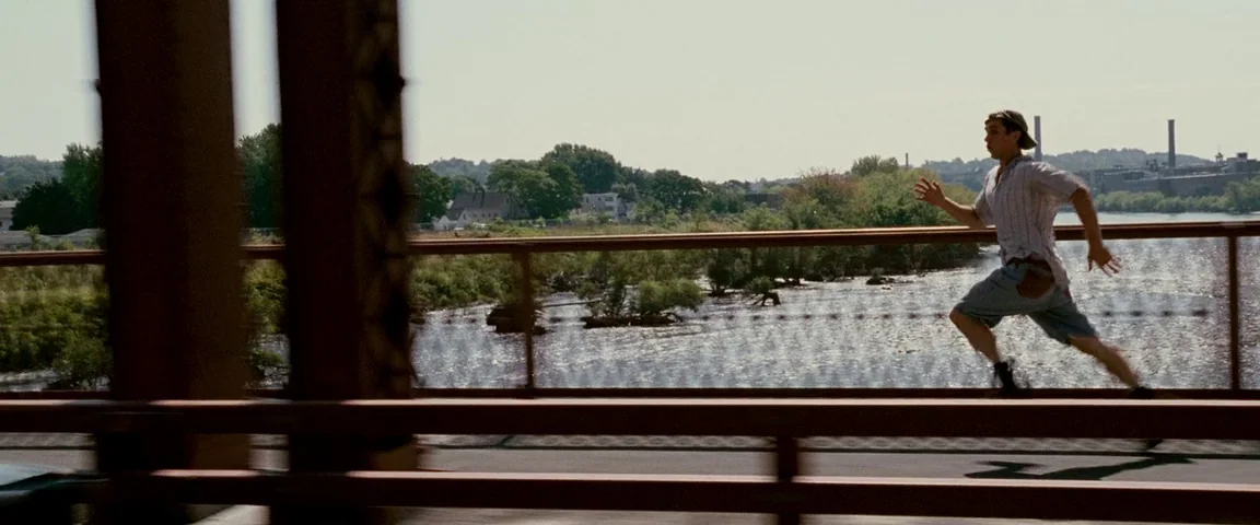The Fighter (2010) movie still: fullbody — An outdoor metal bridge overlooks a wide river with trees and industrial chimneys nearby. A young man…