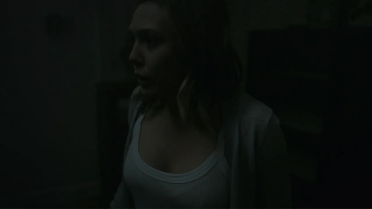 Silent House (2011) movie still: medium — A dimly lit interior room at night with deep shadows surrounding the subject. A woman in a white tank top…