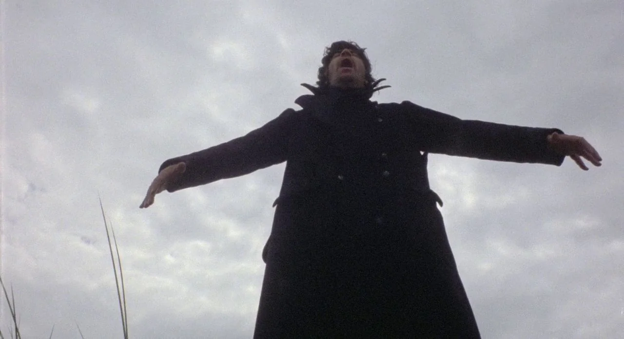The Shout (1978) movie still: fullbody — An open field under an overcast, grey sky with sparse grass stalks visible. A man in a dark overcoat stands…