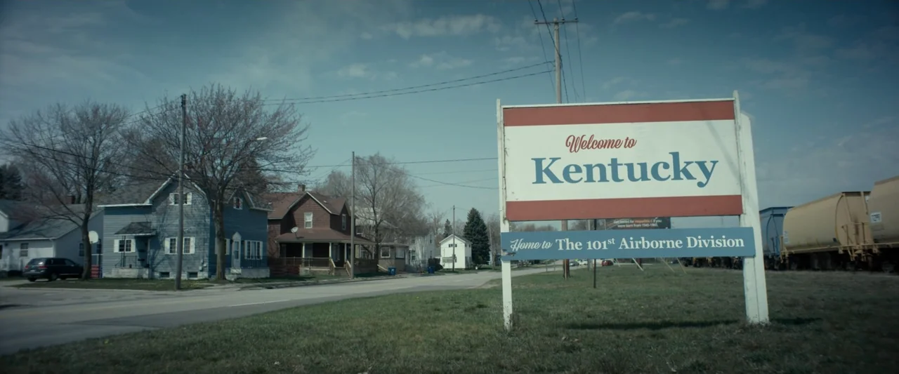 God Bless the Broken Road (2018) movie still: establishing — A suburban roadside scene features a large welcome sign standing in a grassy area.…