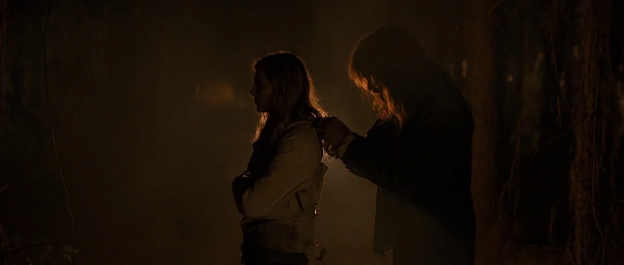The East (2013) movie still: medium — A dark, atmospheric woodland setting at night obscured by heavy shadows. A man stands behind a woman fastening…