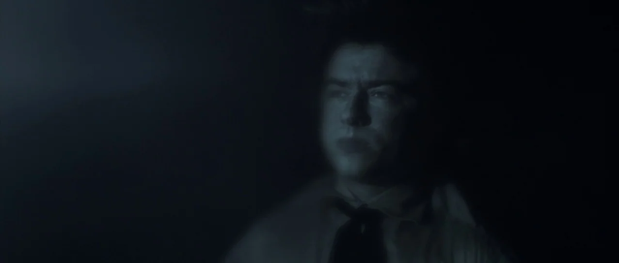 Vanishing Waves (2012) movie still: closeup — A dimly lit room filled with deep shadows obscuring the surroundings. A young man sits in the…