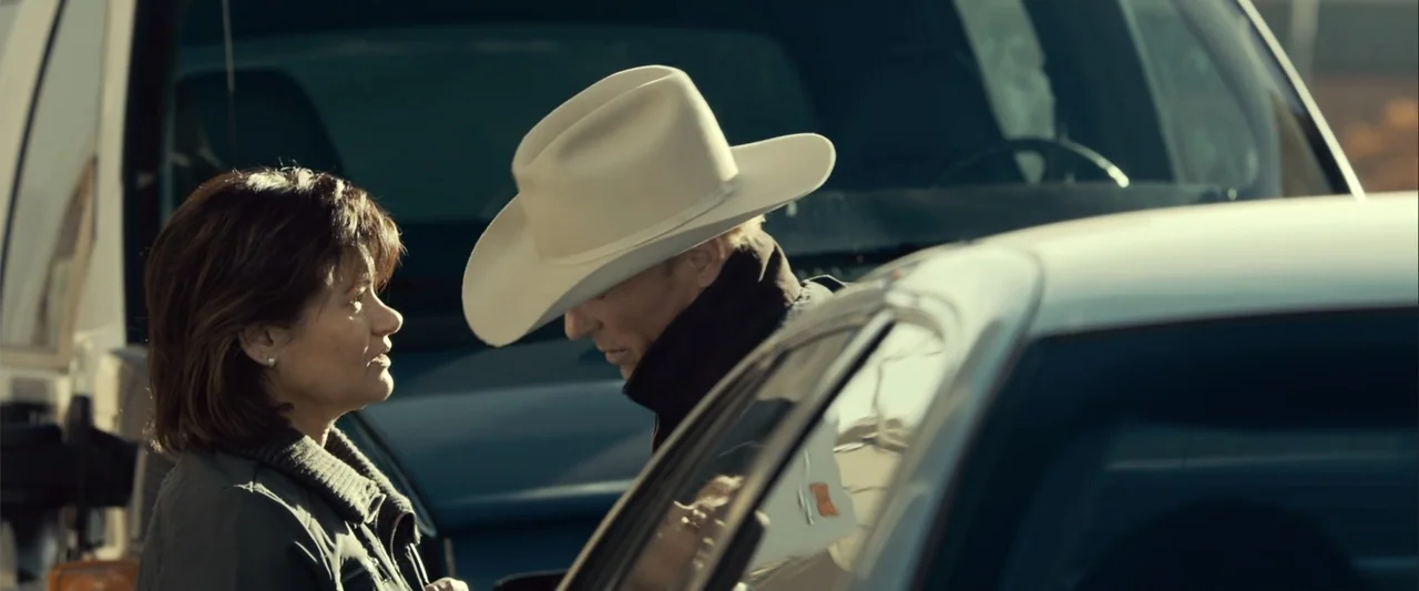 Frontera (2014) movie still: medium — A man and woman interact near parked vehicles in an outdoor lot. The woman faces the man while the man looks…