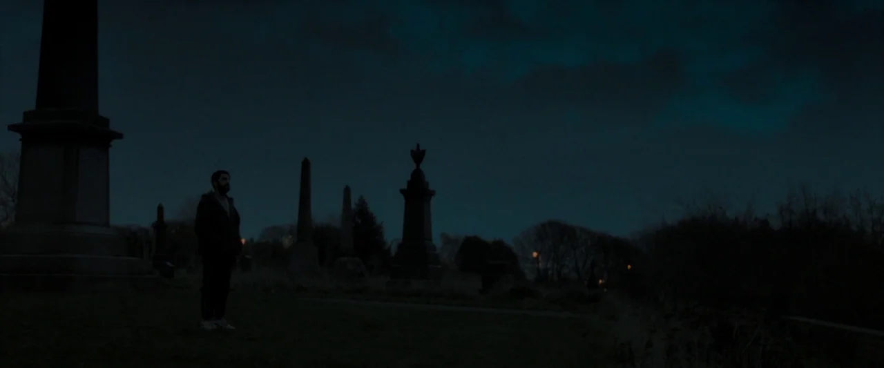 Ali & Ava (2021) movie still: wide — A dark cemetery at night is filled with stone obelisks and gravestones under moonlight. A solitary man stands…