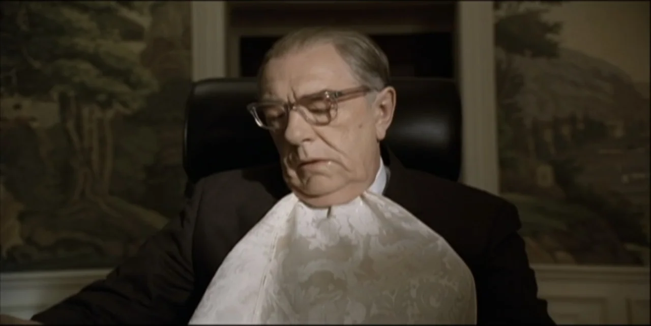 Path to War (2003) movie still: medium — An interior space features wallpaper with a landscape pattern and a dark leather chair. An elderly man with…