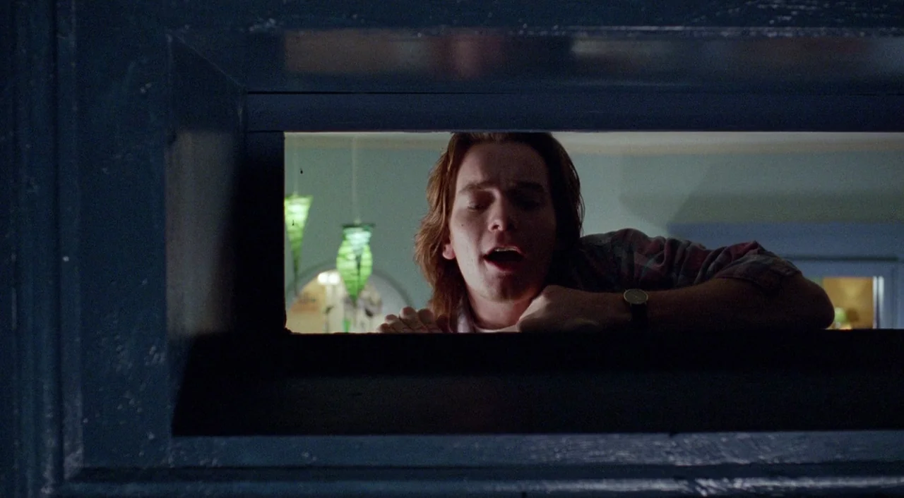 Shallow Grave (1994) movie still: medium — A dark frame of a doorway surrounds a man leaning into a brightly lit space. The interior beyond the door…