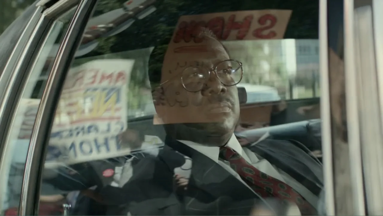 Confirmation (2016) movie still: medium — A man sits in the backseat of a car with protest signs visible through the window. The interior of the…