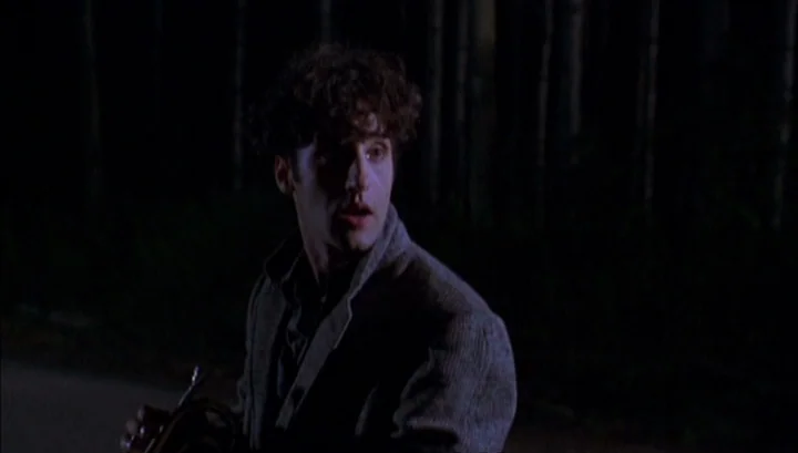 Highway 61 (1991) movie still: medium — A man stands in front of a dense, dark forest at night. The man holds a brass instrument and looks off to…
