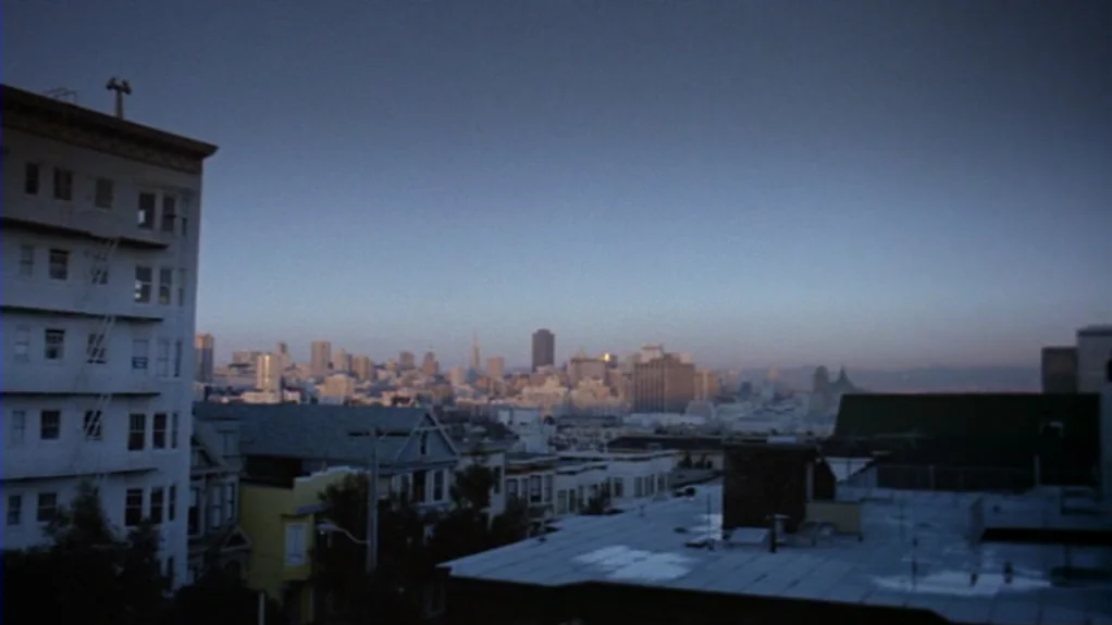 Patty Hearst (1988) movie still: establishing — An elevated view shows a densely packed city skyline under a clear evening sky. Low rooftops of…