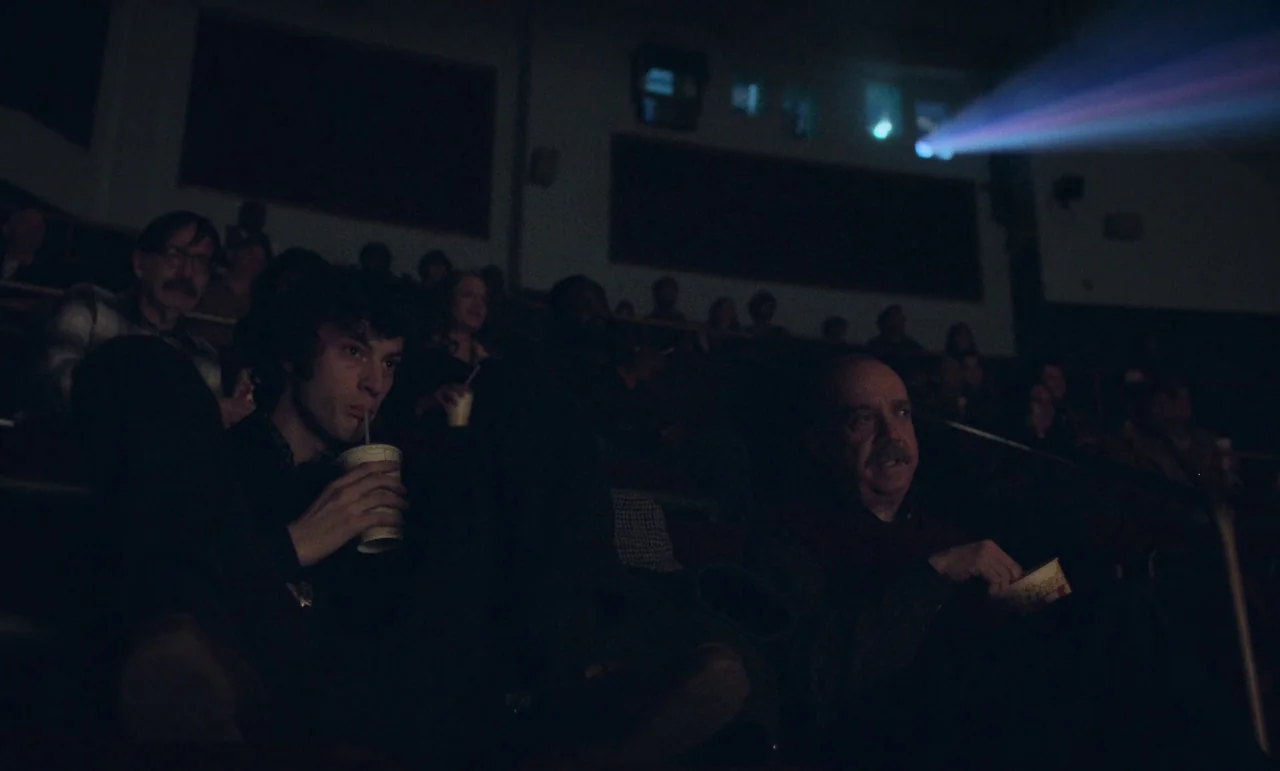 The Holdovers (2023) movie still: medium — A dark movie theater with audience members sitting in rows of tiered seats. Two men in the foreground…