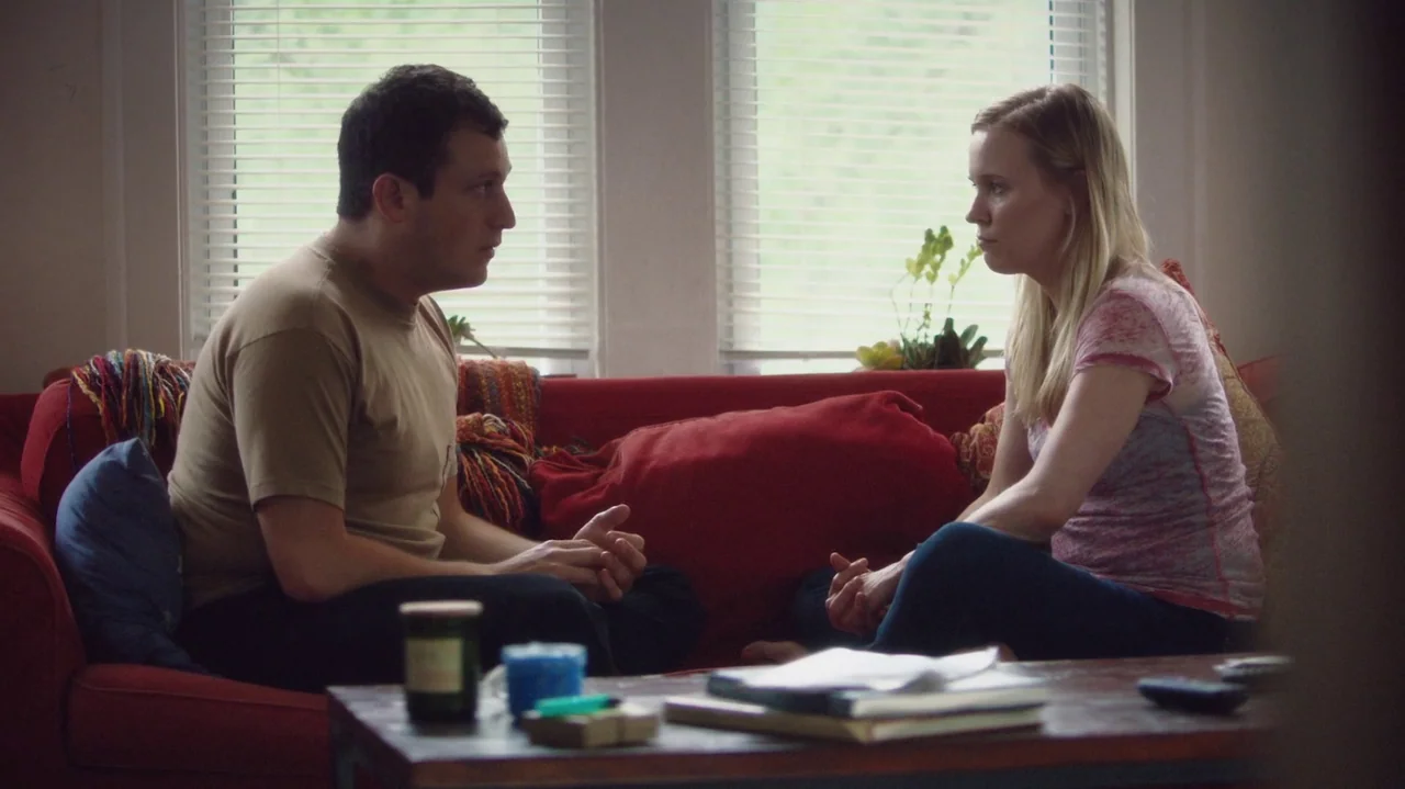 Saint Frances (2019) movie still: medium — A man and a woman sit on a red couch in front of a window. They face each other while engaged in…