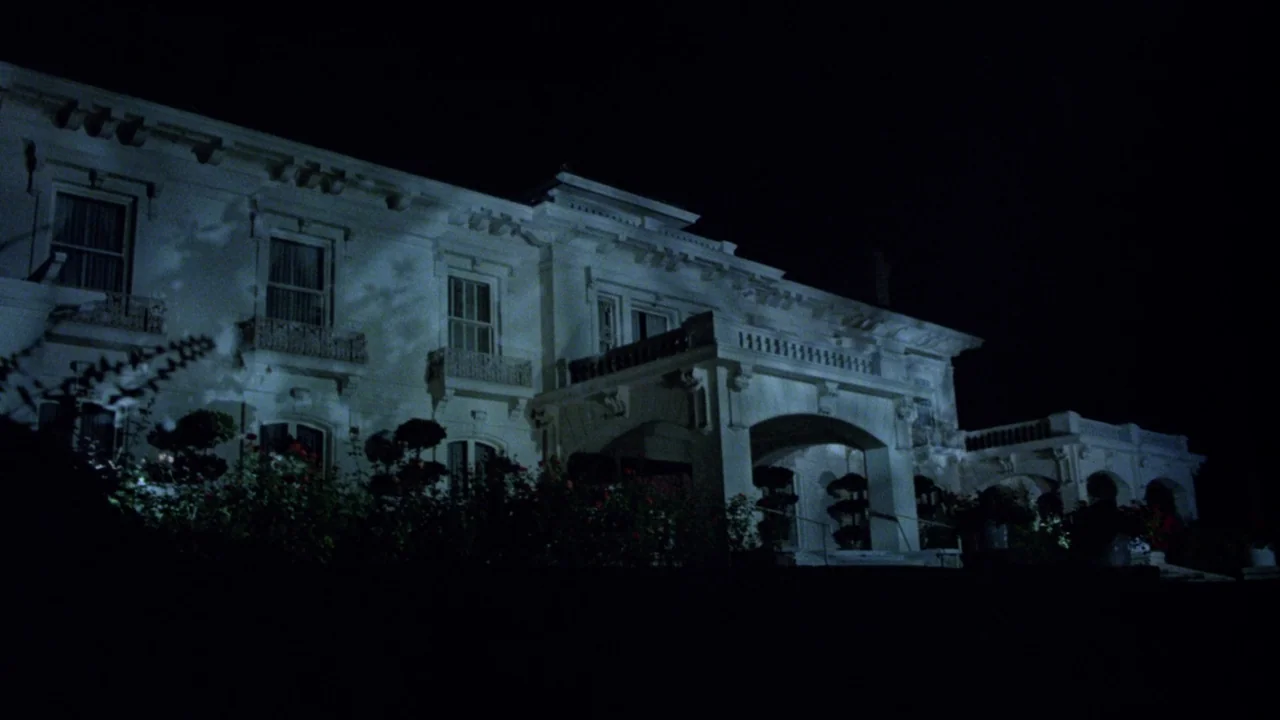 Society (1989) movie still: establishing — A grand white mansion stands prominently against a pitch-black night sky. The building exterior is…