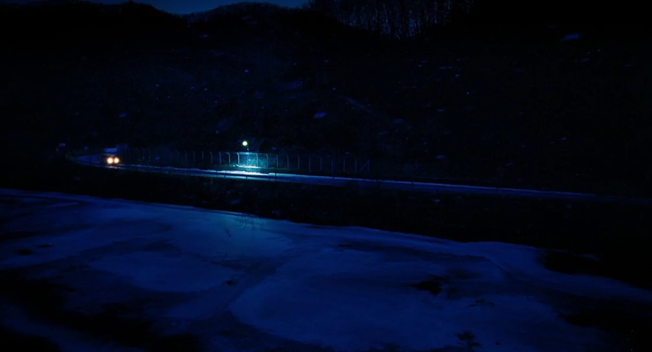 I Saw The Devil (2010) movie still: establishing — A dark road curves through a landscape dominated by a frozen, ice-covered field. A single vehicle…