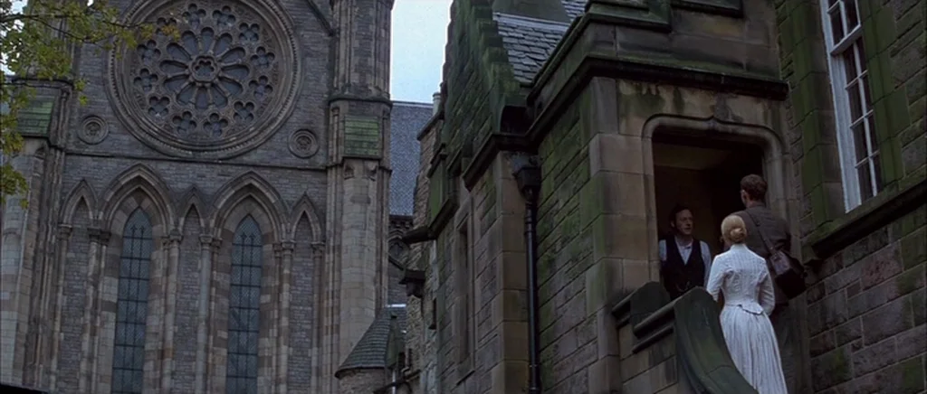 Jude (1996) movie still: wide — A gothic stone cathedral dominates the frame next to a smaller stone building entry. Three people stand gathered at…