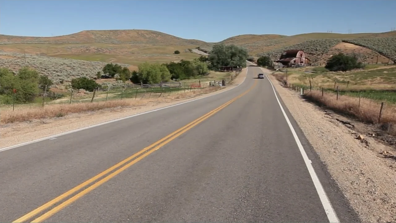Idaho: The Movie (2012) movie still: wide — A desolate two-lane highway winds through arid, sun-baked hills under a clear sky. A single car drives…