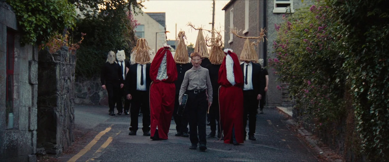 Fréwaka (2024) movie still: fullbody — A group of figures wearing straw masks walks down a paved residential road. A man leads the procession while…