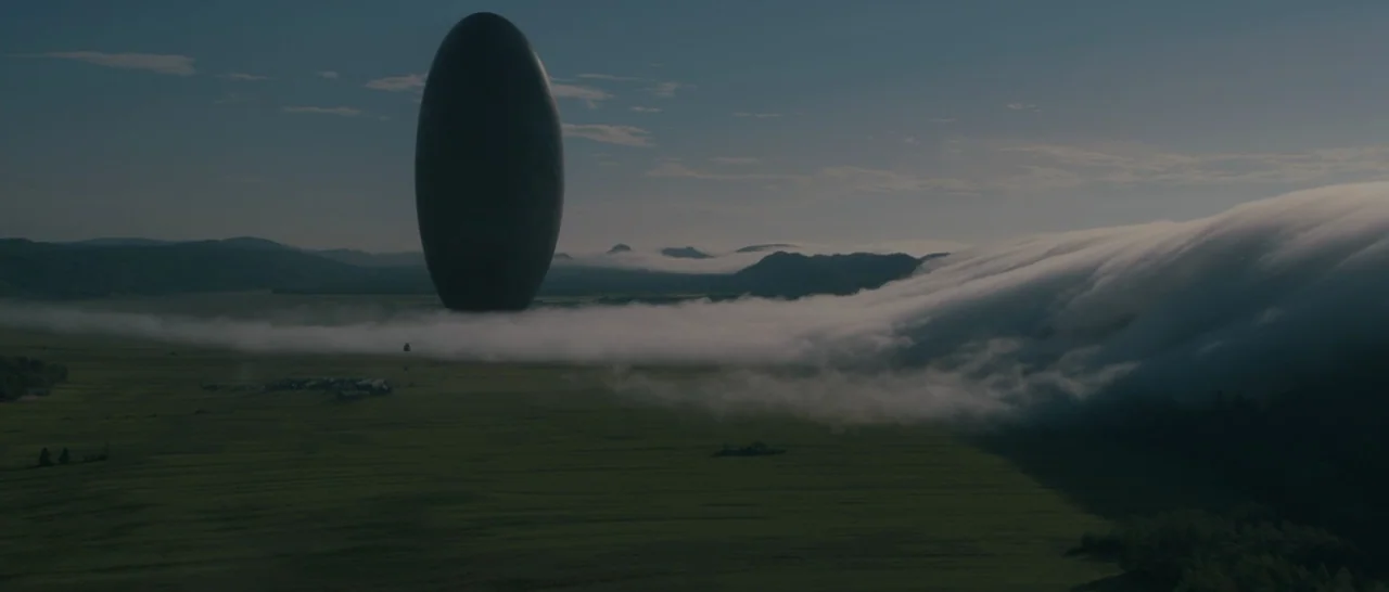 Arrival (2016) movie still: establishing — A vast green field is occupied by a single, monolithic, dark, egg-shaped structure. A low-lying band of…