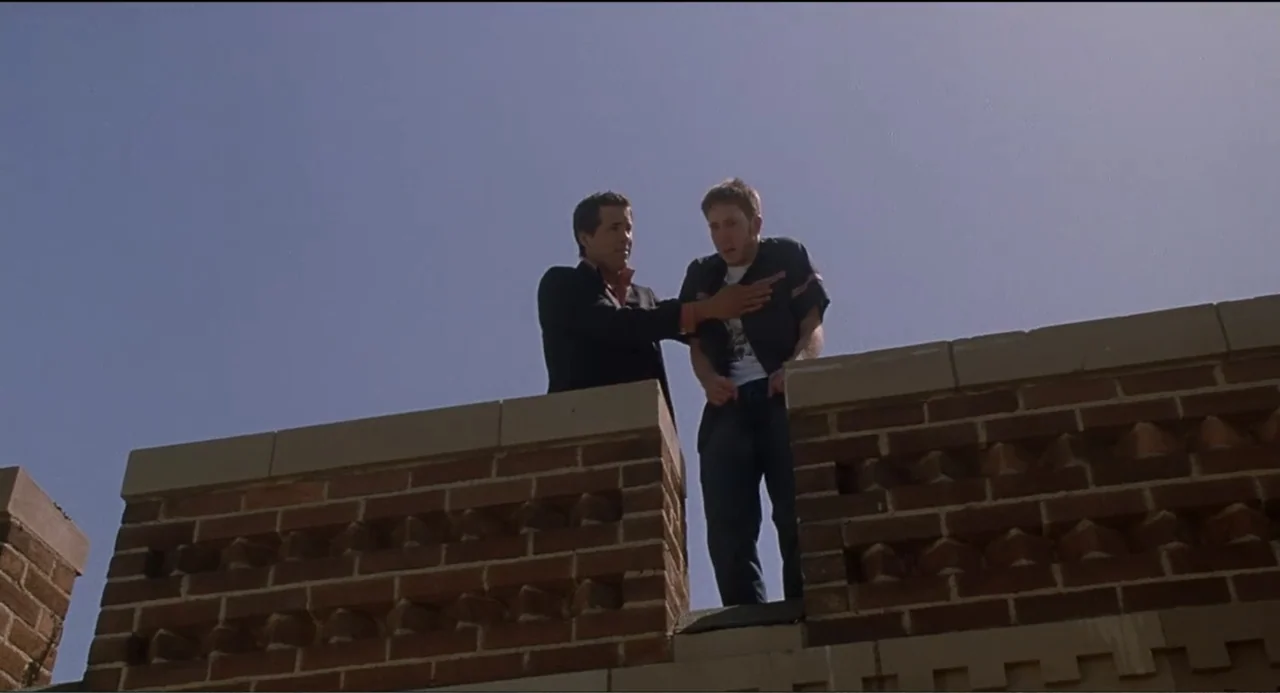 National Lampoon's Van Wilder (2002) movie still: medium — Two men stand atop a weathered red brick wall against a clear blue sky. One man reaches…