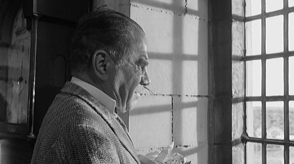 Cul-de-sac (1966) movie still: medium — An interior space features rough-hewn stone walls and a tall, barred window. A man in a blazer stands before…