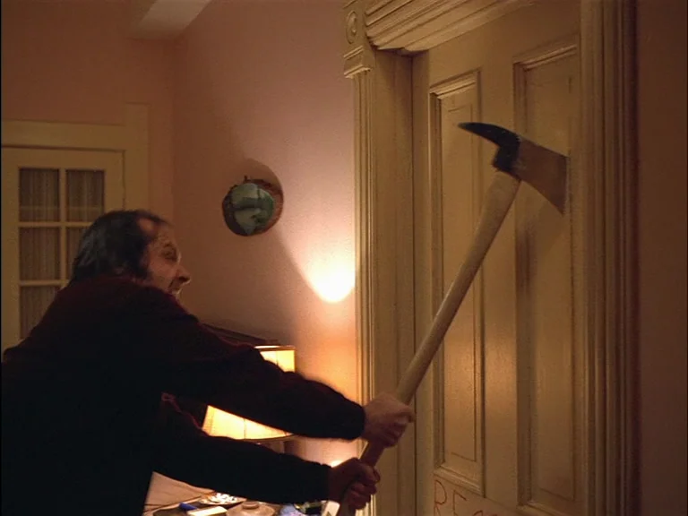 The Shining (1980) movie still: medium — An interior hallway features a light-colored wooden door and a wall-mounted picture. A man in a dark shirt…