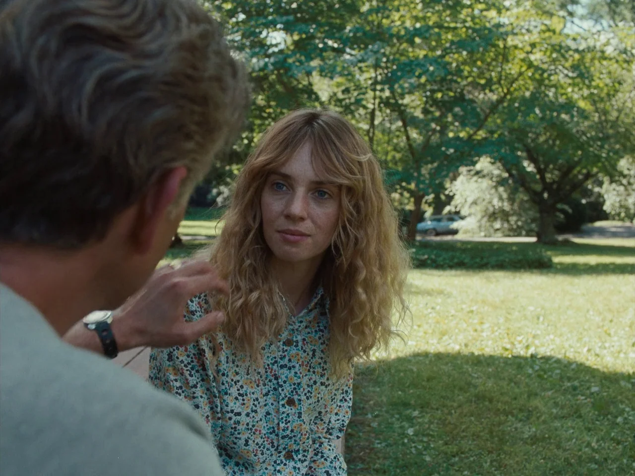 Maestro (2023) movie still: medium — A sunny park with large deciduous trees and a grassy lawn in the background. A woman in a floral shirt sits…